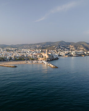 Aerial view of the Church of Sant Bartomeu i Santa Tecla overlooking the Mediterranean Sea, sandy beaches, and white coastal buildings in Sitges, Catalonia, Spain.