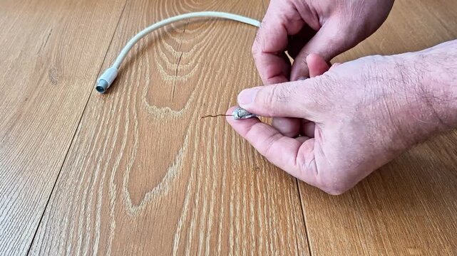 Hands holding the exposed copper wire end of a coaxial cable on a wooden surface, demonstrating preparation or repair process for electrical and signal connections