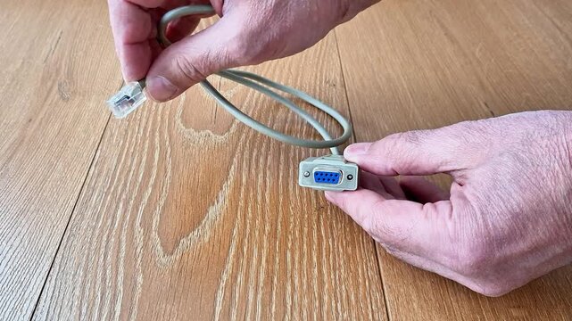 A grey cable with an RJ45 connector and a DB9 female serial port held against a wooden background. Detailed view of hardware interface and connectivity components for data transfer