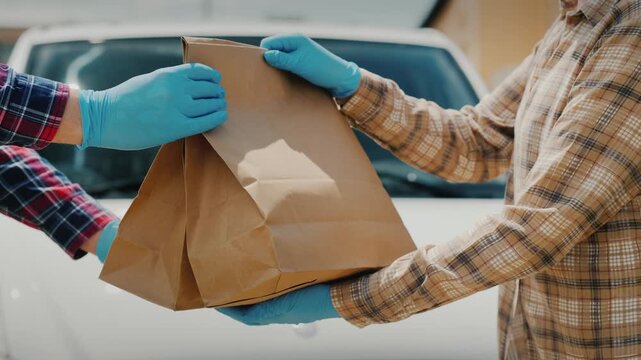 Courier in protective gloves handing over paper bags with food to customer against white delivery van.