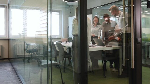 Group of business people standing together and discussing their work and projects, having a team meeting in an office, view from glass wall