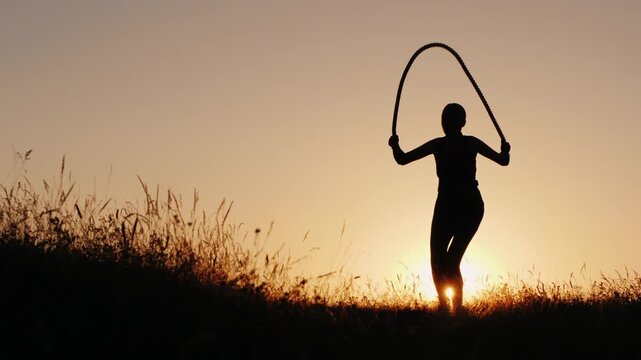 Silhouette of active woman jumping rope at sunset in field with golden sky and glowing sun rays.