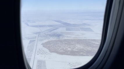 Fototapeta premium Airplane window view over snowy landscape and frozen fields, soft winter light and calm atmosphere, travel perspective suggesting quiet journey and remote terrain
