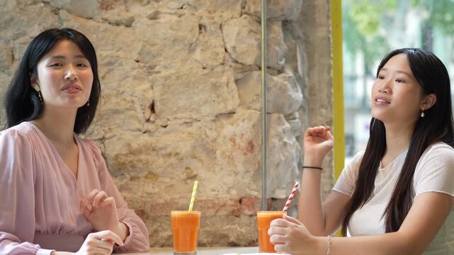 Two women laugh at a cafe table. One holds her hand to her ear. The other smiles, eyes closed. Orange drinks sit on the table. Stone walls and glass divide the space