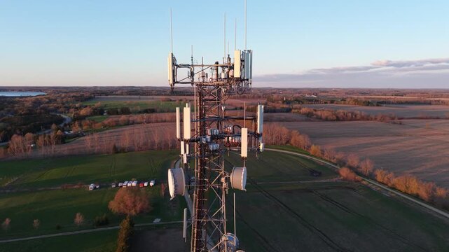 A cinematic aerial drone pull-back from a lattice telecommunications tower at golden hour, revealing a commercial warehouse facility and expansive agricultural land under a bright sunset