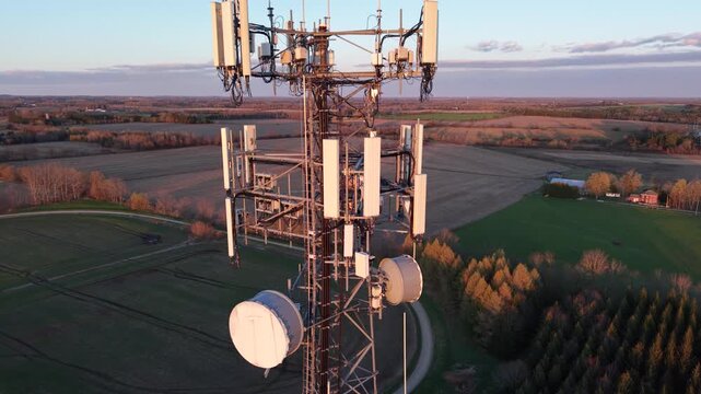 A smooth aerial drone descent alongside a steel lattice telecommunications tower at golden hour, detailing cellular sector antennas and vast agricultural fields