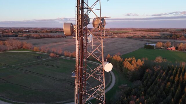 A close-up aerial drone ascent alongside a steel lattice telecommunications tower, detailing sector antenna arrays and circular microwave drum dishes at sunset