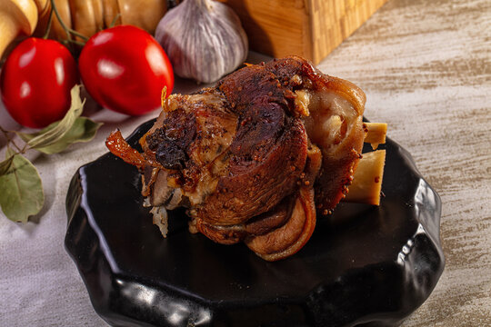 Roasted pork knuckle with crispy skin on a black plate with tomatoes and garlic background