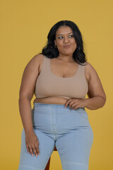 Obraz premium Woman posing on low stool in studio mustard backdrop croptop jeans hoops adjusting waist