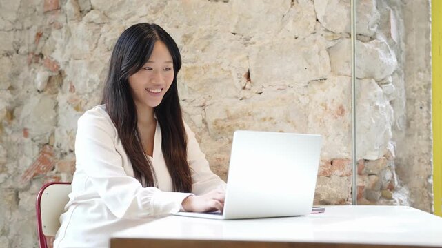 Young woman smiles while typing on laptop. Light stone wall background adds rustic charm. She appears focused, happy, and engaged. Ideal for business, education, or remote work themes