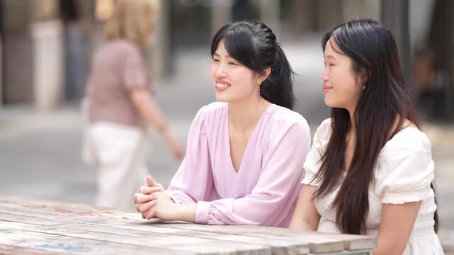 Two women sit at a wooden table outdoors. One smiles warmly, eyes directed forward. The other looks toward her, listening attentively. Soft daylight frames their relaxed, friendly interaction