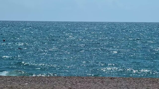 Sunlit turquoise sea pebbled beach, gentle waves shimmer under clear sky, empty shoreline, wide panoramic frame conveying tranquil