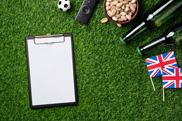 Naklejka premium Blank clipboard, TV remote, bowl of pistachios, green beer bottles, mini soccer ball and England flags on green grass create a perfect game viewing setup. Top view with copy space.