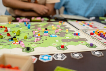 Fototapeta premium Close-up of people playing a complex strategy board game with colorful wooden pieces and hexagonal tiles, representing logical thinking, resource management, and social gathering at home or cafe.
