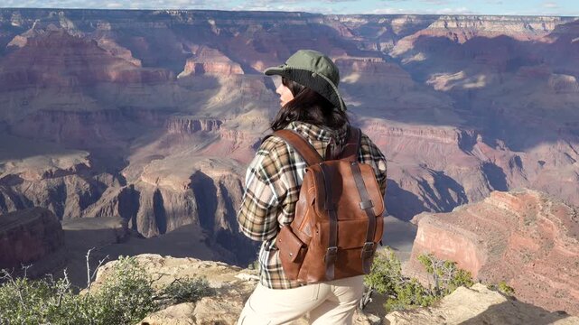 Young female traveler with a backpack enjoying the beautiful panoramic view from the edge of the grand canyon south rim, turning around and smiling at the camera on a sunny day in arizona