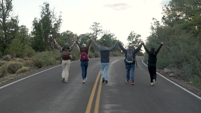 Group of five happy friends with backpacks walking away on an empty road in grand canyon national park, holding hands and raising their arms in celebration of their successful journey