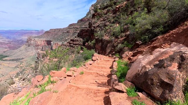 Personal perspective of a hiker descending the narrow, winding switchbacks of the bright angel trail, offering a breathtaking view into the vast expanse of the grand canyon on a sunny day