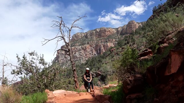 Young female hiker sitting on a rock, taking a break along a dusty path in grand canyon national park, arizona, with impressive red rock canyon walls and blue sky in the background