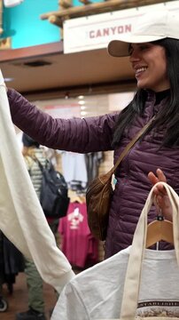 Smiling young woman wearing a baseball cap and puffy jacket browsing through clothes on hangers in a gift shop, looking for the perfect souvenir from her memorable travel experience