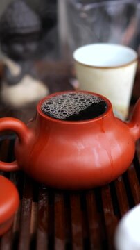 Hot steaming water pouring into a traditional clay teapot with loose leaf tea during an authentic asian tea ceremony, creating a relaxing and mindful beverage experience on a wooden tray