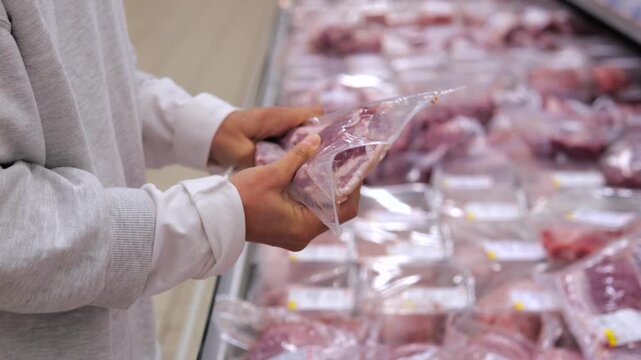 Unrecognizable customer in a supermarket choosing a piece of vacuum sealed raw pork belly from the refrigerated display case, carefully inspecting the quality before buying it for cooking