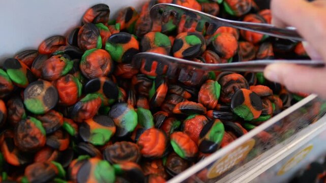 Close up view of a person using metal tongs to pick up spooky pumpkin shaped licorice gummies from a bulk bin in a candy store, a festive treat for a halloween party or trick or treating