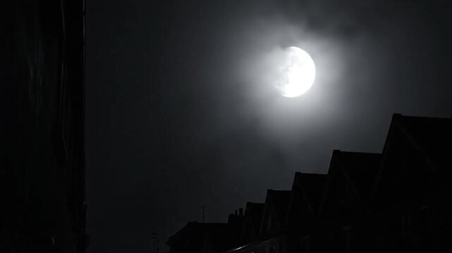 Eerie nighttime scene featuring a glowing full moon partially obscured by clouds above a row of silhouetted houses