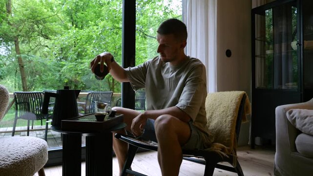 Focused man sitting indoors and enjoying a traditional chinese tea ceremony, pouring hot pu erh tea from a chahai into a small cup with a lush green forest visible through a large window