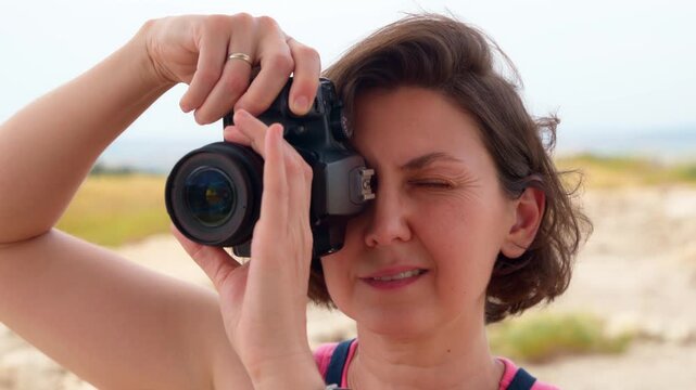 Close up of a focused middle aged female photographer holding a professional DSLR camera and looking through the viewfinder. Woman taking photos outdoors during a bright day in a natural setting