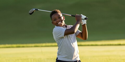 Naklejka premium Young African american boy swings golf club on green course. Child enjoys outdoor sport game, smiles with happiness, focused on hitting ball. Healthy active pastime for kids.