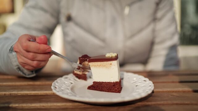 Woman hands taking piece of chocolate layered cake with fork on white plate in outdoor cafe.