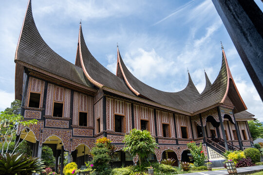 Wide angle low perspective of traditional Rumah Gadang Minangkabau, showcasing dramatic curved roof and rich ornamental wooden facade under blue sky