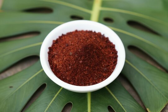 Red annatto seeds powder in white bowl on green leaf. Natural coloring agent for food and paint. Tropical spice ingredient from Bixa orellana plant.