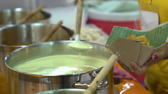Vendor serving hot curry dish in paper box with banana leaf, close-up Indonesian street food preparation at food stall