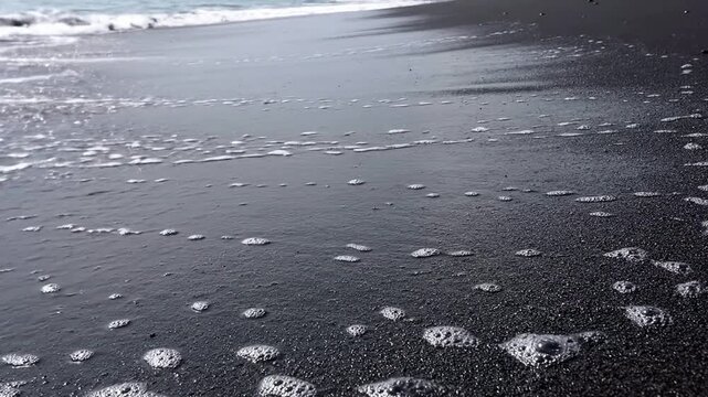 Gentle ocean waves washing over dark volcanic sand beach, creating foam bubbles, natural texture, serene coastal scene