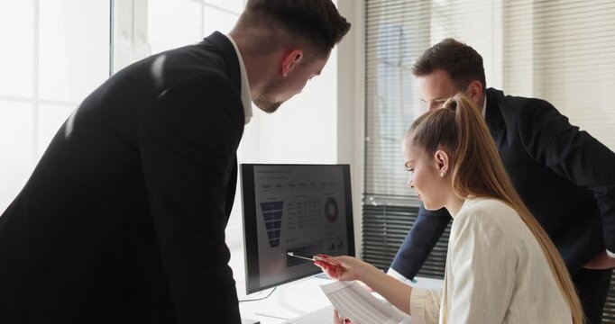 Team colleagues meeting in office for analysis. Analysts point to charts on computer, compare documents, discuss results, refine planning. Strong business planning and teamwork concept.