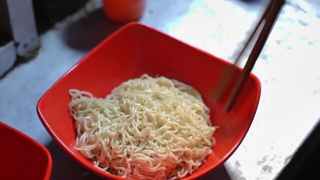 Bakmi noodles being mixed with sauce in a red bowl, close-up Indonesian noodle preparation with chopsticks at a food stall