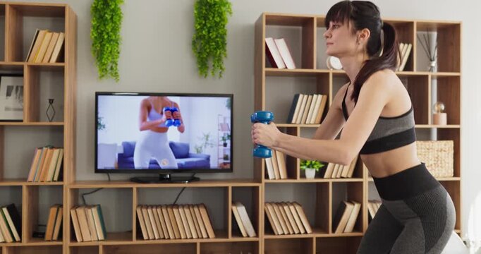 Woman home workout with dumbbells via online television. She squats in a living room, following a fitness training video and building strength, balance and tone. Concept of remote home fitness.