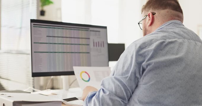 Man accountant in office doing finance analysis. He checks charts on a computer and reviews paperwork to track budget accuracy and expenses. Clear concept of a stressed manager at work.
