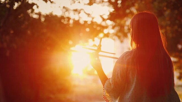 Young woman holding toy airplane against golden sunset sky in nature park.