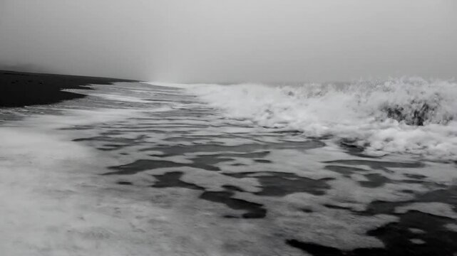 Black sand beach with foamy waves under a hazy grey sky, a monochrome coastal scene