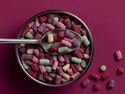 Overhead bowl of pastel cereal pieces with a metal spoon on a deep magenta background. Great for colorful snack packaging, breakfast ads, and social media food design.