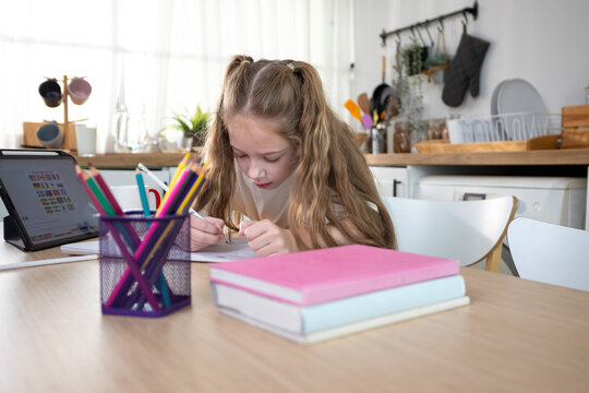 Young girl studying and doing homework at home with digital tablet. Child education, online learning, homeschooling, creativity, and focused learning in a modern kitchen.