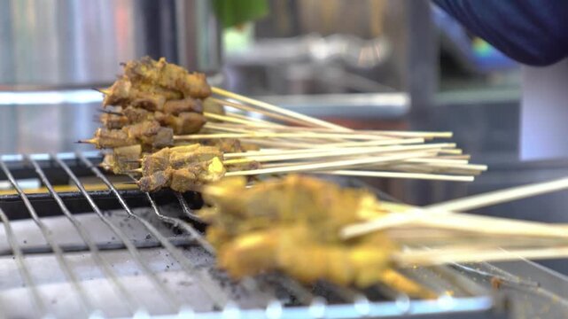 Grilled satay skewers cooking on metal grill, close-up Asian street food barbecue with shallow focus and warm market lighting