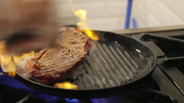 Raw beef steak searing on hot grill pan with flames and smoke, close-up restaurant kitchen cooking preparation