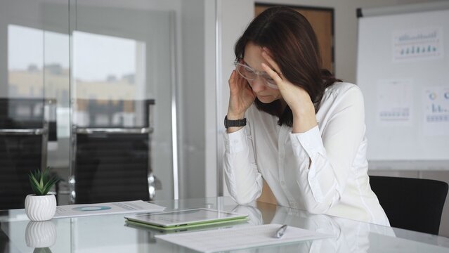 Businesswoman in white blouse experiencing severe headache, feeling overwhelmed by work stress and burnout, holding temples with hands while struggling with mental pressure at office desk