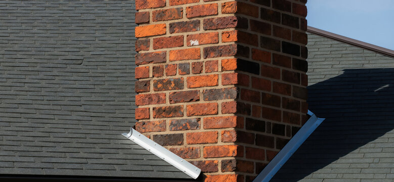 Close-up of a red brick chimney and dark grey shingle roof with metal flashing detail. Sunlight casts a shadow on the roof surface. Residential construction exterior.