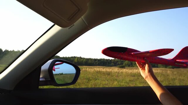 Hand holds red airplane near car window. Sunlit field stretches beyond windshield. Reflections show airplane and trees clearly. Child's hand holds toy with care and delight