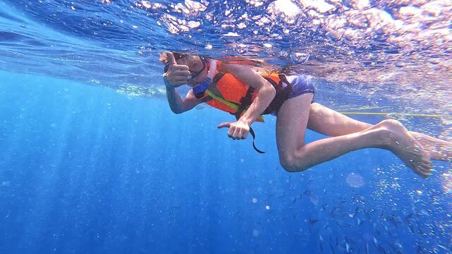 Underwater explorer floats with vibrant orange buoyancy aid. Blue water sparkles with sunlight piercing through surface. Person glides gently, arms extended, eyes focused forward