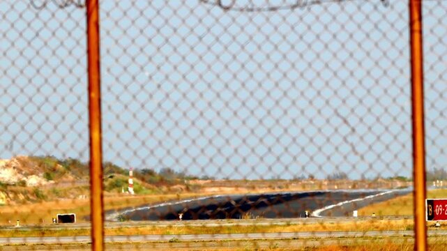 Chain link fence frames a runway beyond. Dirt and grass surround the paved path. Barbed wire adds a sense of restricted access. Sunlight casts warm tones over the scene